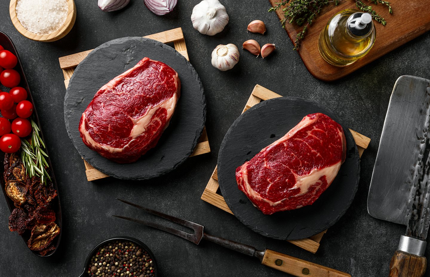 Two raw beef steaks with ingredients and utensils on slate.