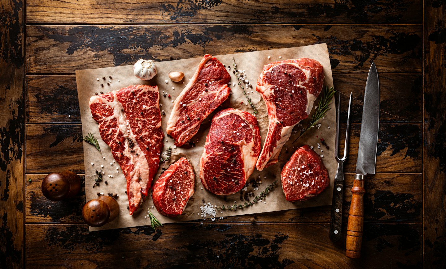 Raw steaks with herbs and garlic on a wooden table.